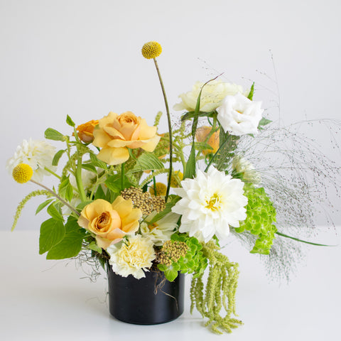 Soft yellow roses and white flowers are arranged in a black vase with billy balls.
