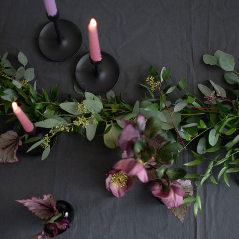 Greenery is spread across a black table cloth. There are also tapered pink and purple candles in black candle holders.