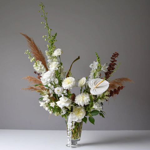 A large flower arrangement in a footed glass vessel. The arrangement is mostly white with hints of brown and greenery.