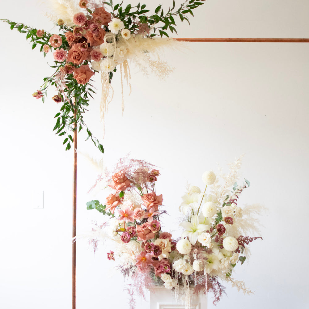 A close-up of the top left corner of a simple copper arch. There are muted blush and cream flowers attached to the poles.
