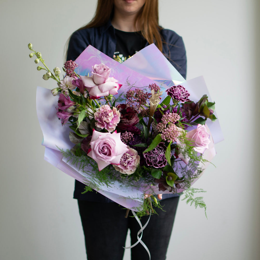 A hand-tied bouquet of various purple flowers wrapped in pretty, shiny paper.