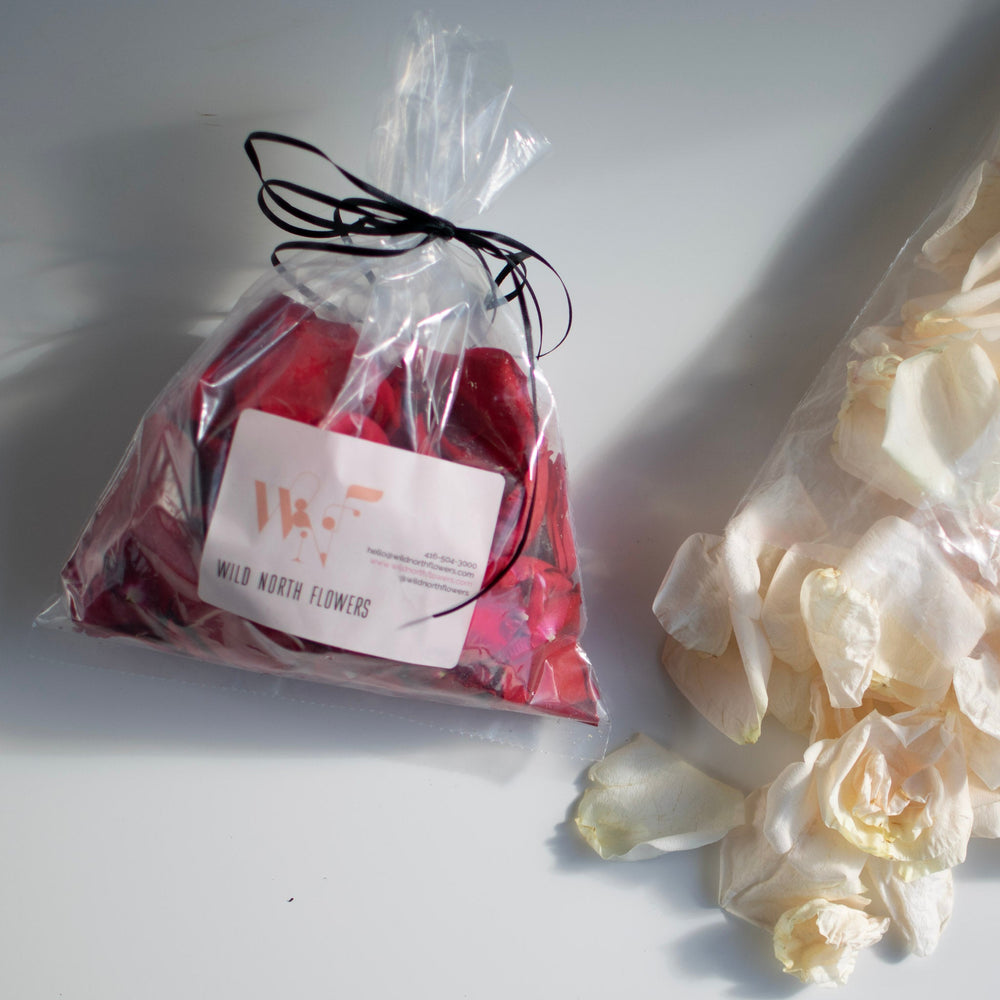 A closed bag of red rose petals lays flat on a white surface. White rose petals are scattered to the right.