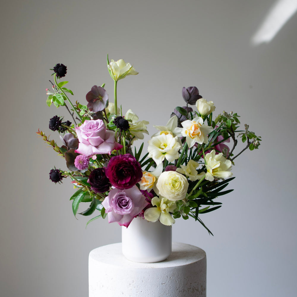 A large, moody flower bouquet in a white vase. The palette is light purple roses, white lilies, and pops of deep purple.