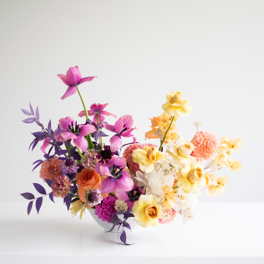 A two-toned flower arrangement with rich purples and delicate yellows in a white bowl.