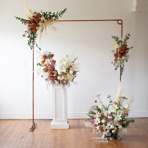 A copper arch with flowers brackets a large floral arrangement on a white pedestal. Another large arrangement has been placed on the floor in front of the arch.