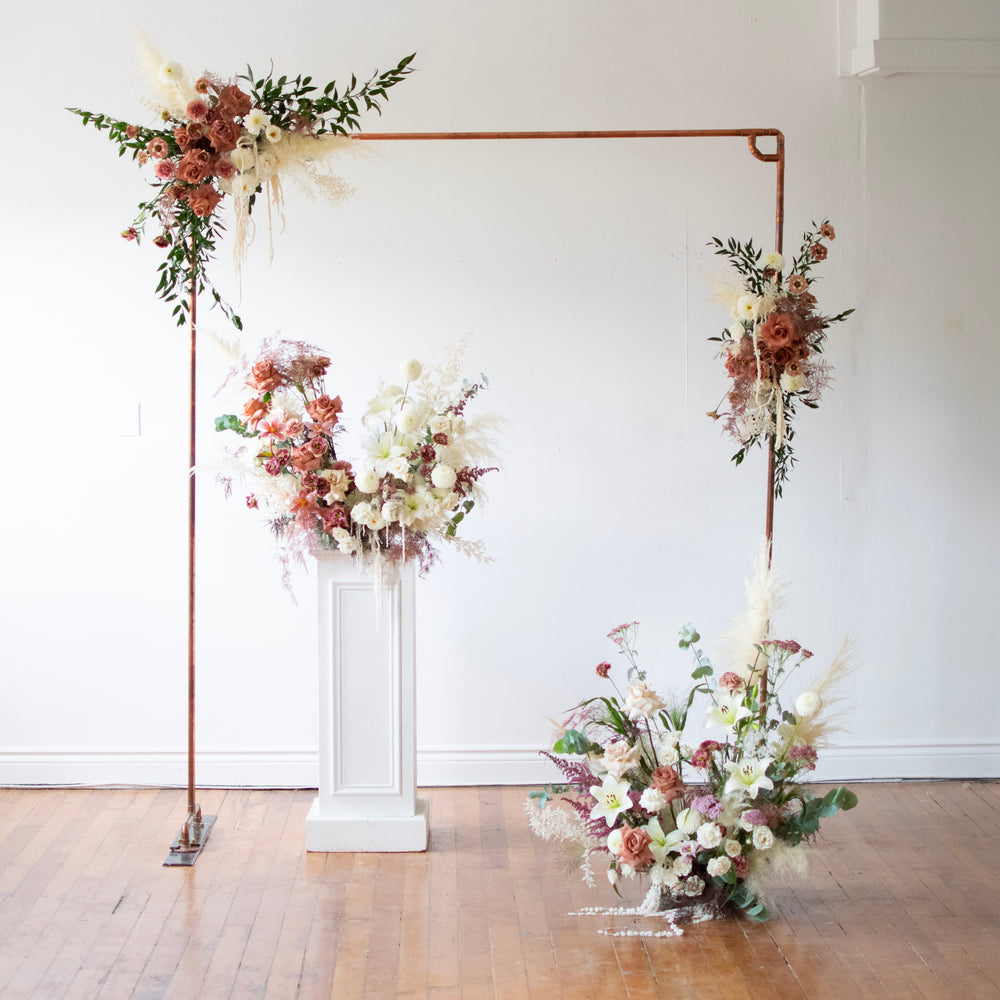 A floor piece accompanies a simple arch and pedestal, all adorned with pink and cream wedding flowers.