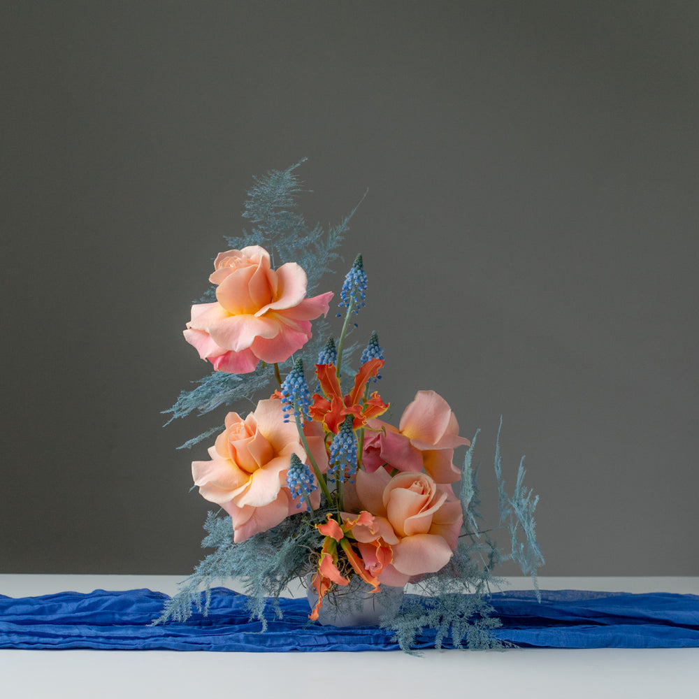 Small floral arrangement with peach roses and blue and orange accents in a small white vase.