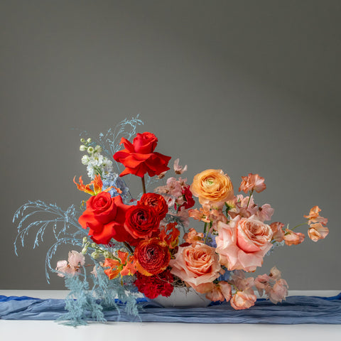 Large vibrant centrepiece with many large blooms in red and peach with blue accents.