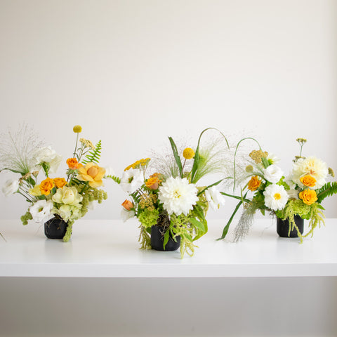 Three mini fresh flower arrangements with white and rich yellow flowers in black vases.