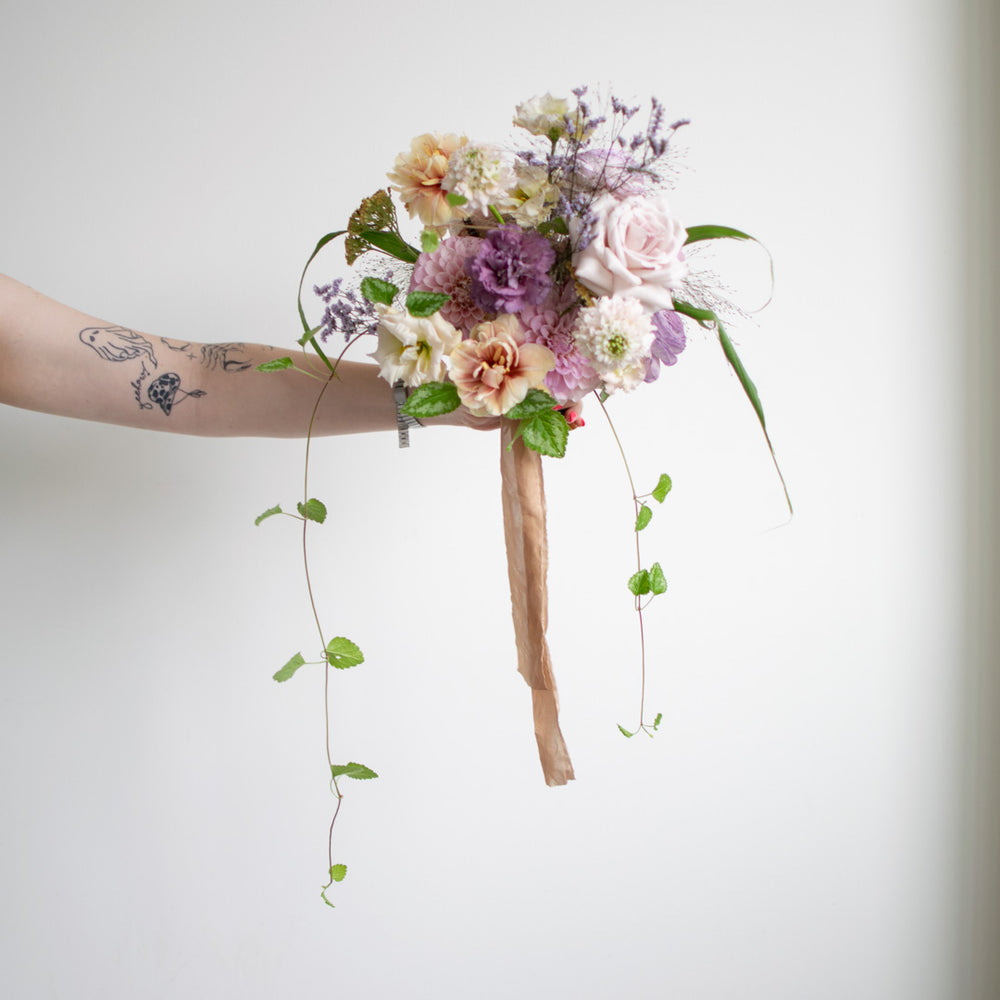 A small, romantic bouquet in soft pastels and neutral accents is wrapped with trailing silk.