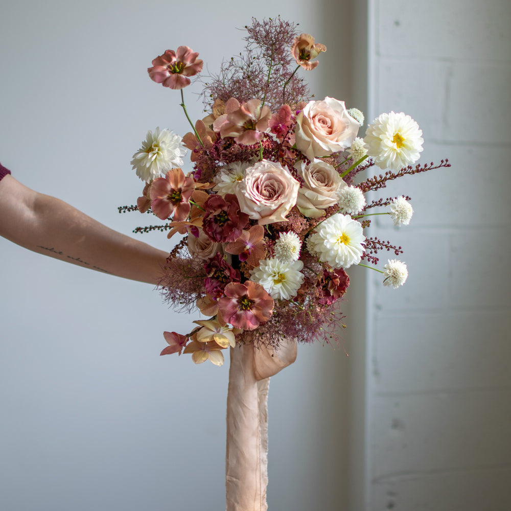 A muted pink bouquet is held out by a forearm. The overall palette is blush tones, but there are pops of white.