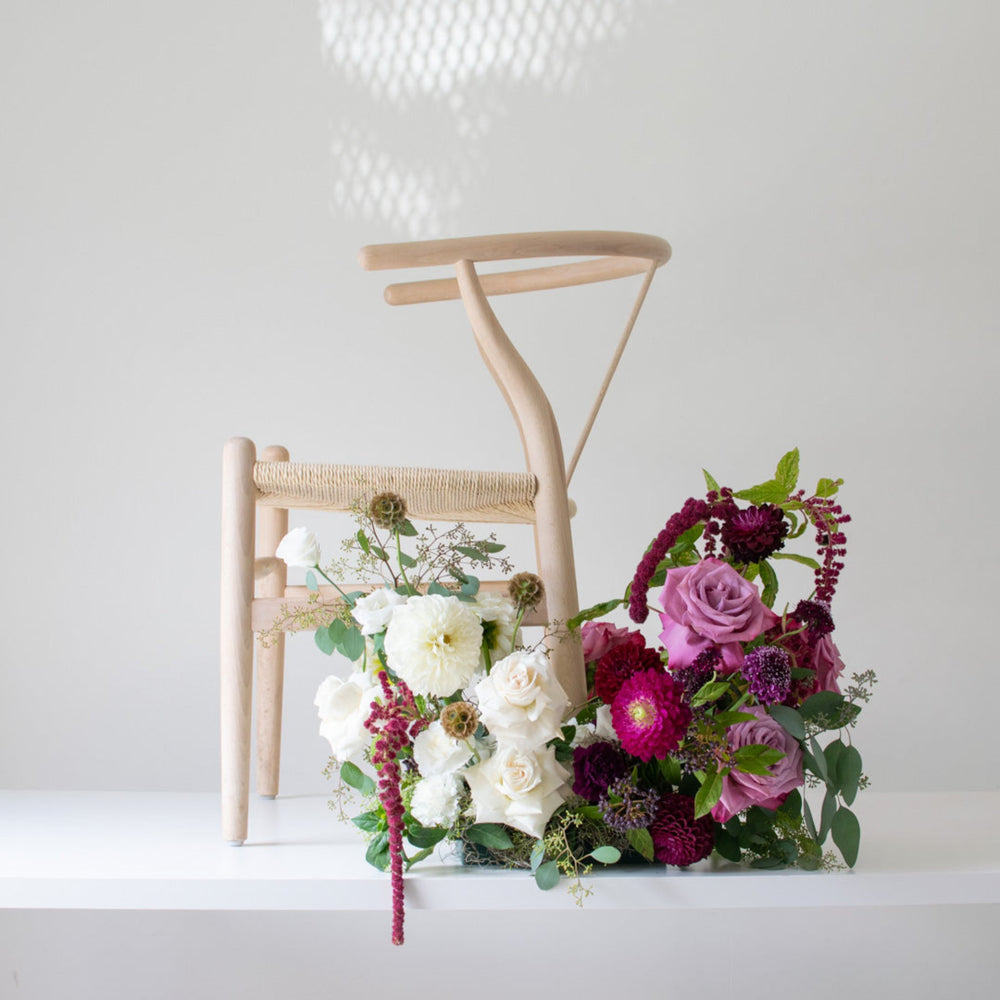 A small floral arrangements sits in front of a wooden chair. The chair is in profile, facing left, and the flowers are deep purple and white.