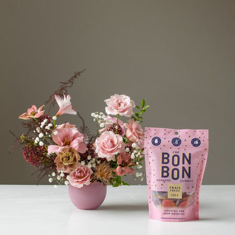 A bag of gourmet candy sits next to a petite floral arrangement with a blush palette.