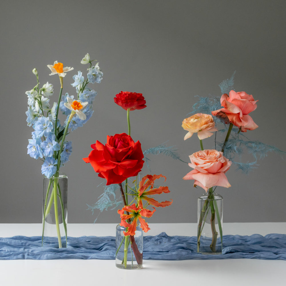 Bright red roses, pink and peach ranunculus, and blue delphinium in glass bud vases.