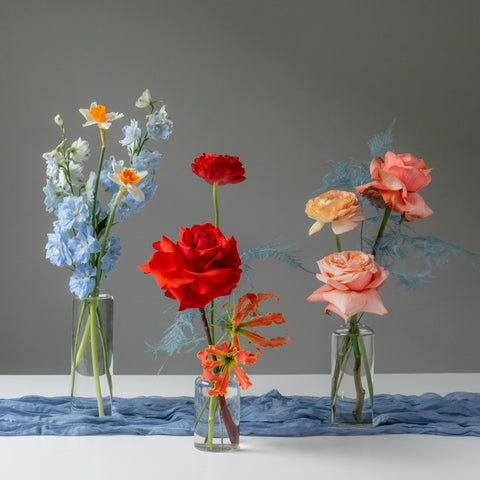 Bright red roses, pink and peach ranunculus, and blue delphinium in glass vases.