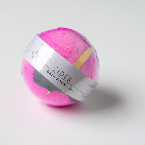 A hot pink bath bomb wrapped in plastic. The grey label says "Cider Bath Bomb - 4oz"