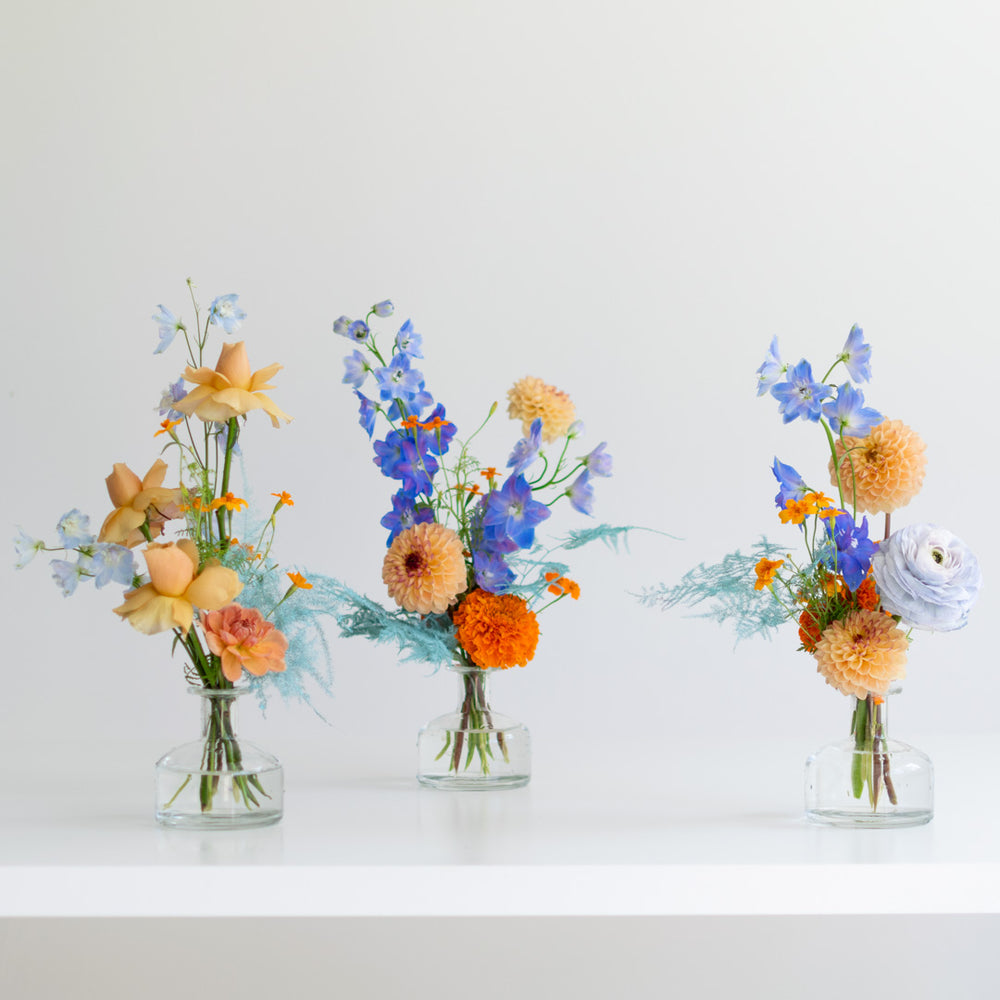 Three mini floral arrangements sit in glass bud vases. The flowers have an orange and blue palette.