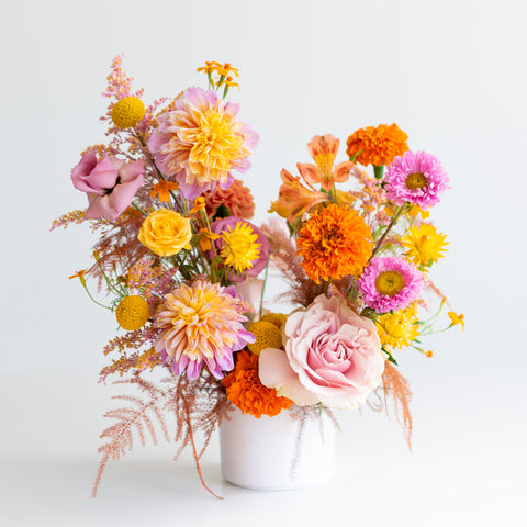 A bright orange, yellow, and pink floral arrangement in a white, cylindrical vase