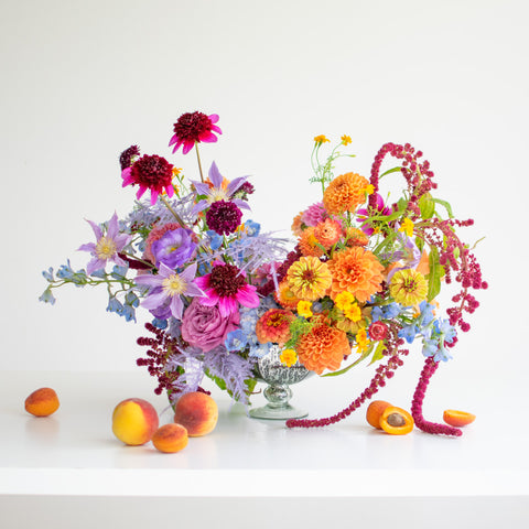 A large two toned floral arrangement with bright oranges and subdued purples in a silver, footed vase. Fruits sit on the table beside it.