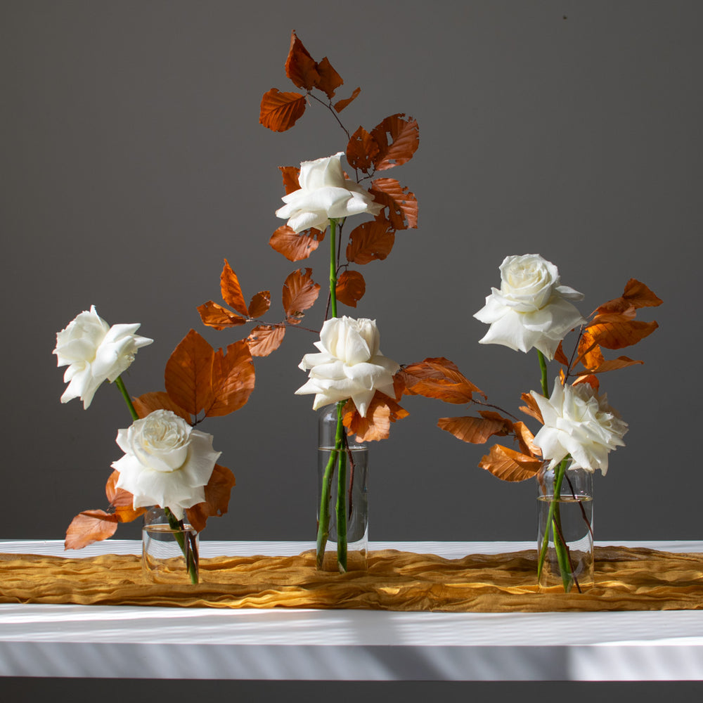 White roses and autumn leaves sit elegantly in a trio of clear bud vases.