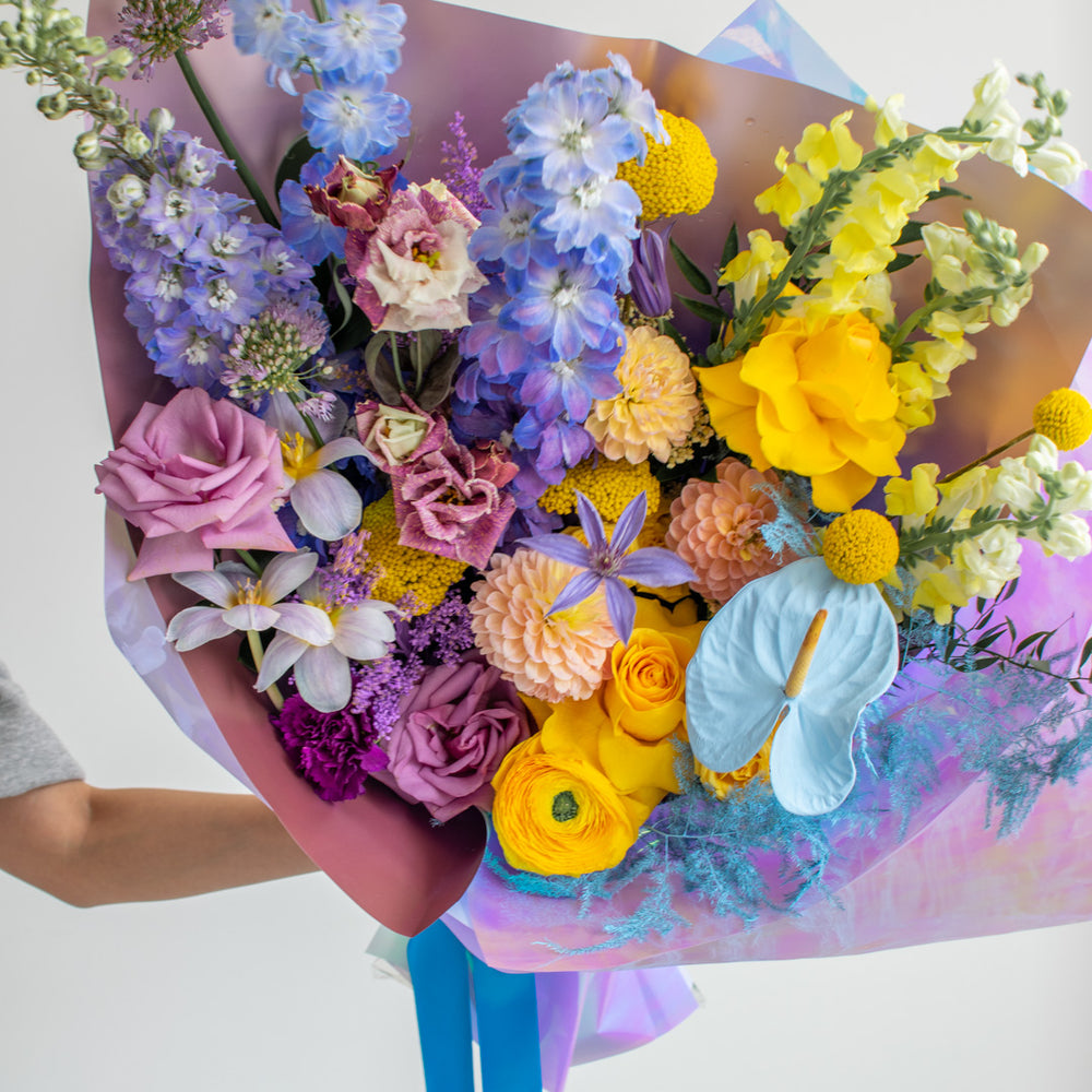 A close-up of bright yellow, purple, and light blue flowers in a hand tied bouquet.