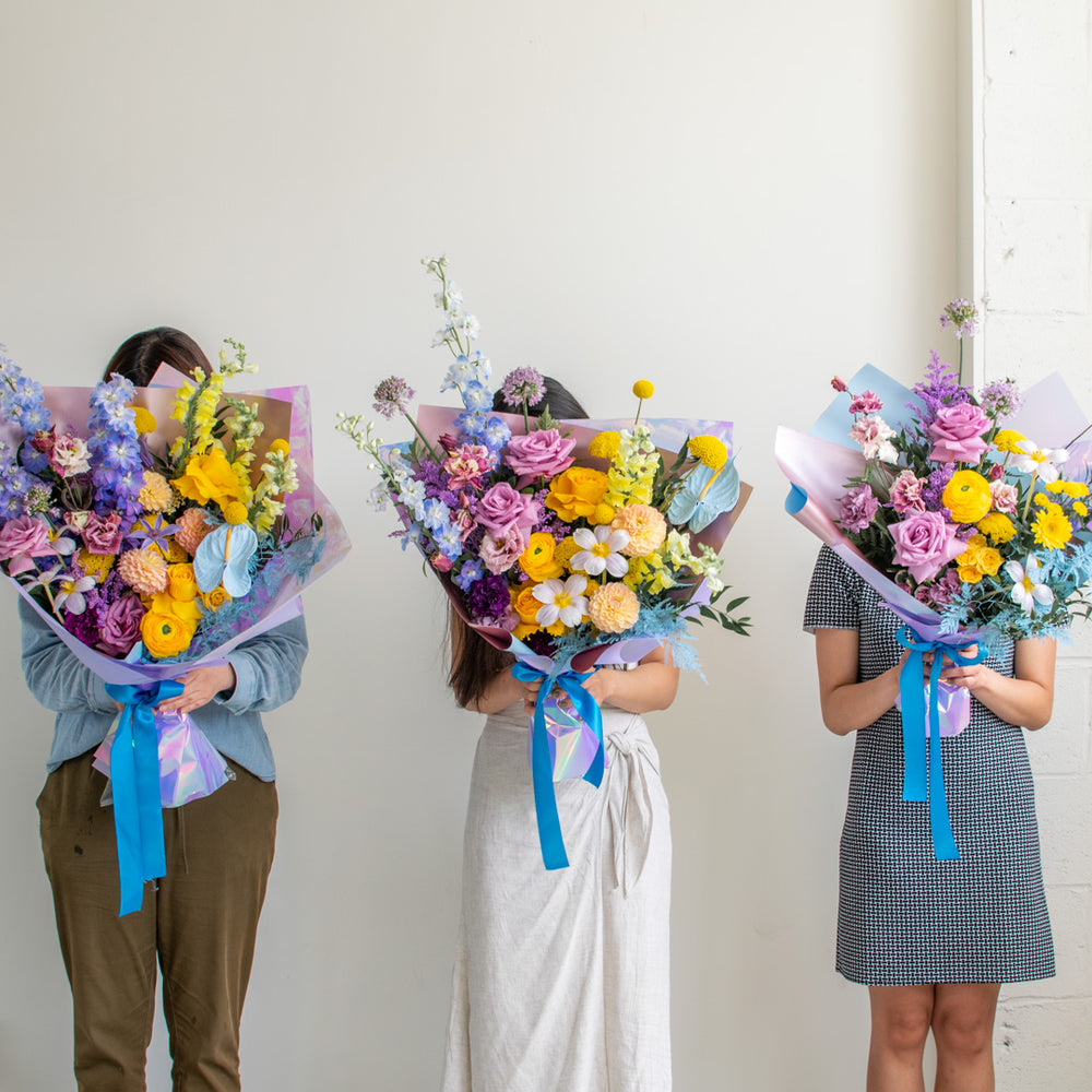 Three women stand together, holding three sizes of fresh flower bouquets.
