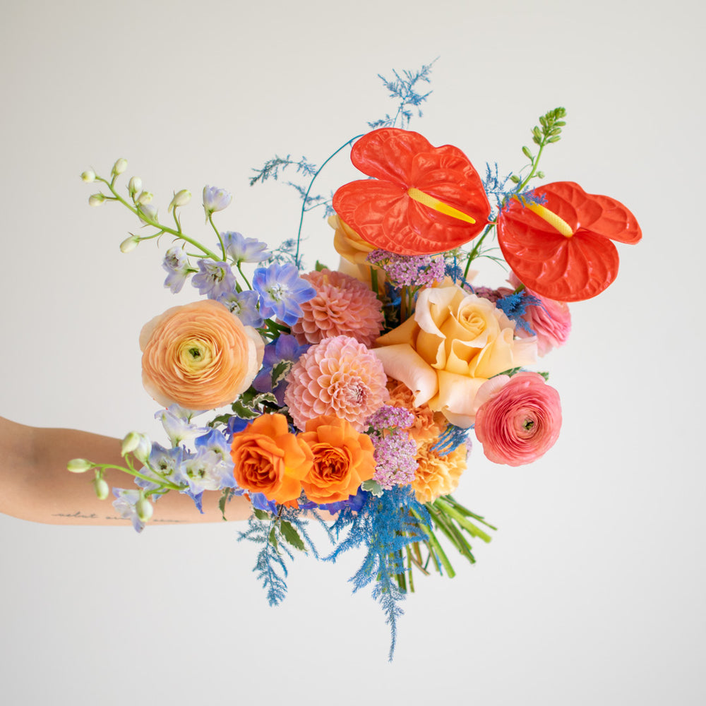A bright and colorful bouquet containing red anthuriums, orange roses, dahlias and ranunculus.