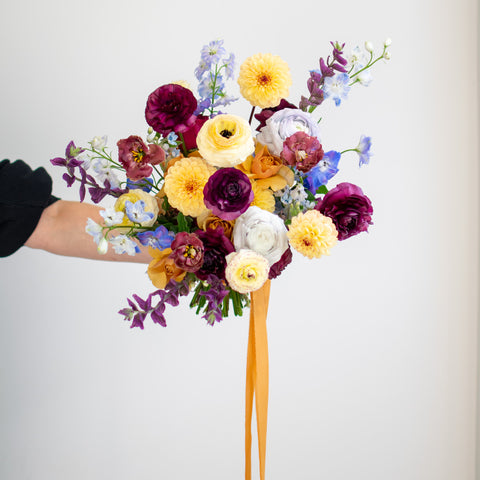 A large bridal bouquet is being held out. There are light blues, deep purples, light yellows, and a trailing orange ribbon.