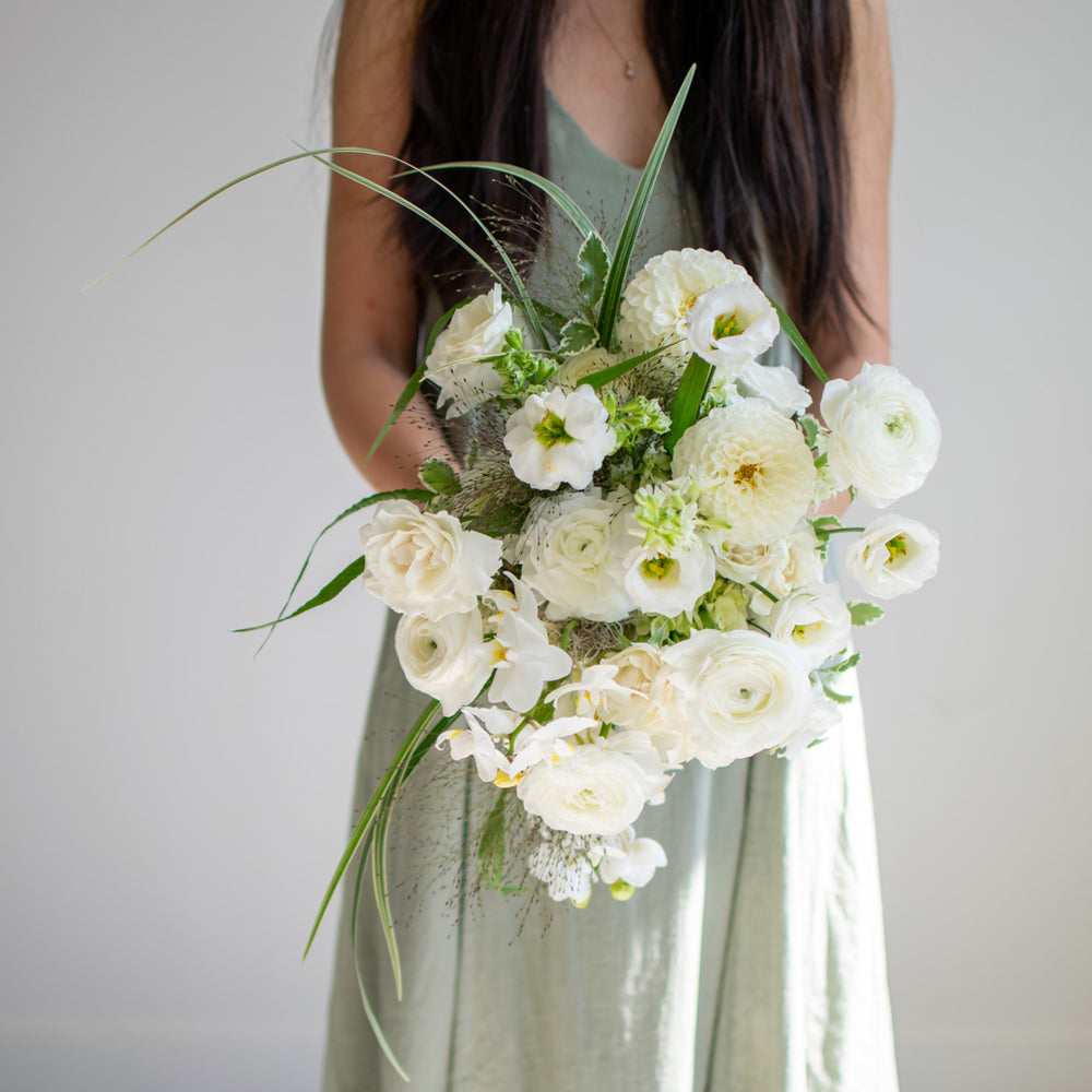 A woman in a sage green dress holds a white bridal bouquet with lots of fresh greenery.