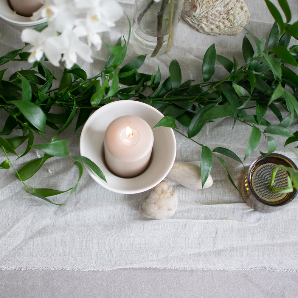 A neutral candle sits in a white dish next to a vibrant, fresh greenery table runner.