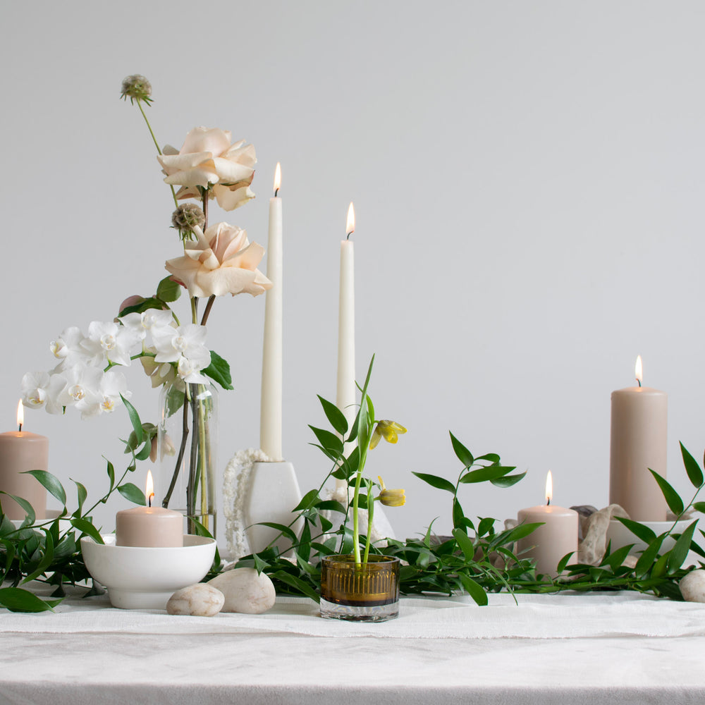 A simple tablescape with light pink and white bud vases, greenery, and many candles.