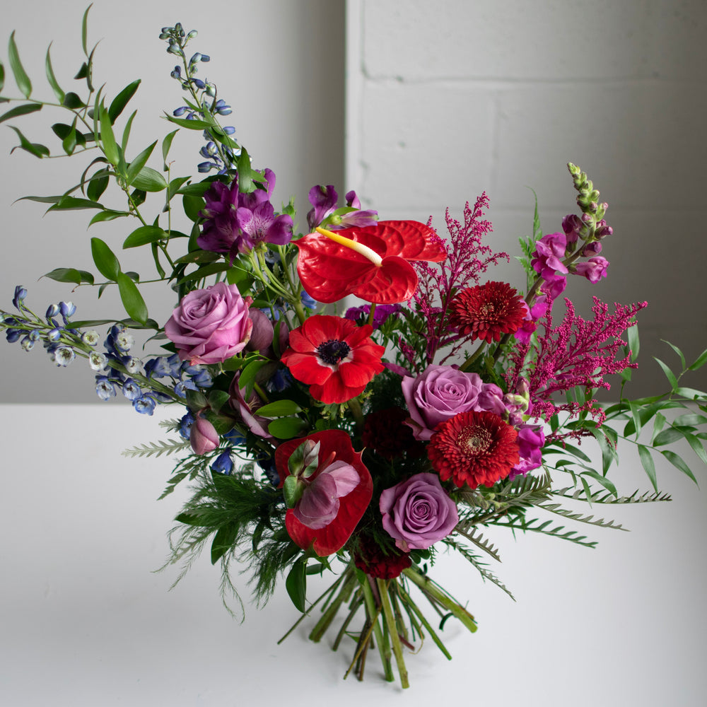 Deep red and purple flowers in a hand-tied bouquet. Roses are daisies are included.