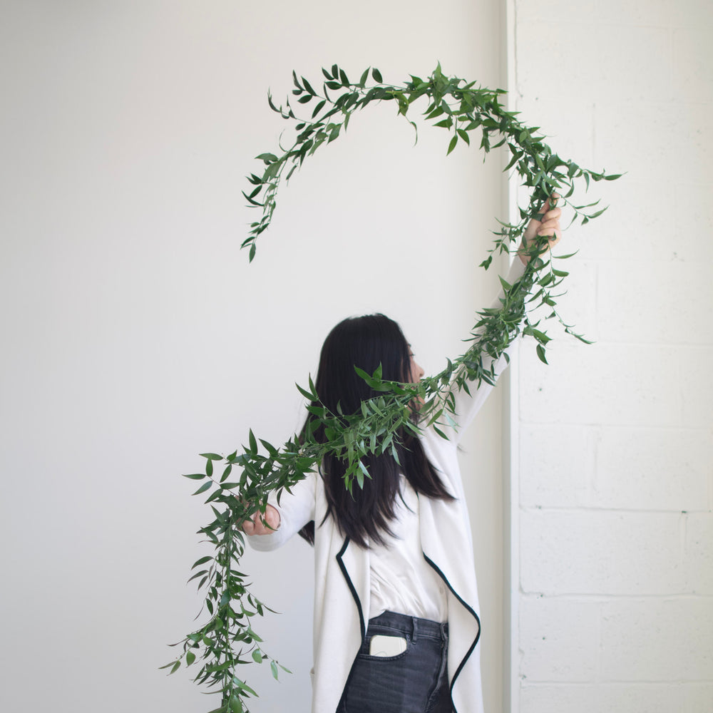 A woman in a white top and dark jeans holds a simple greenery garland over her head, which is cresting.