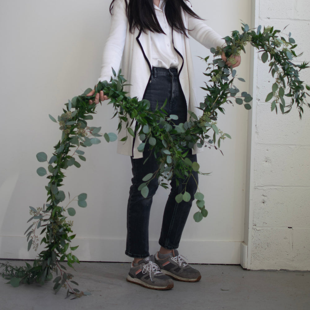 A person in a white coat and blue jeans holds a simple greenery garland.