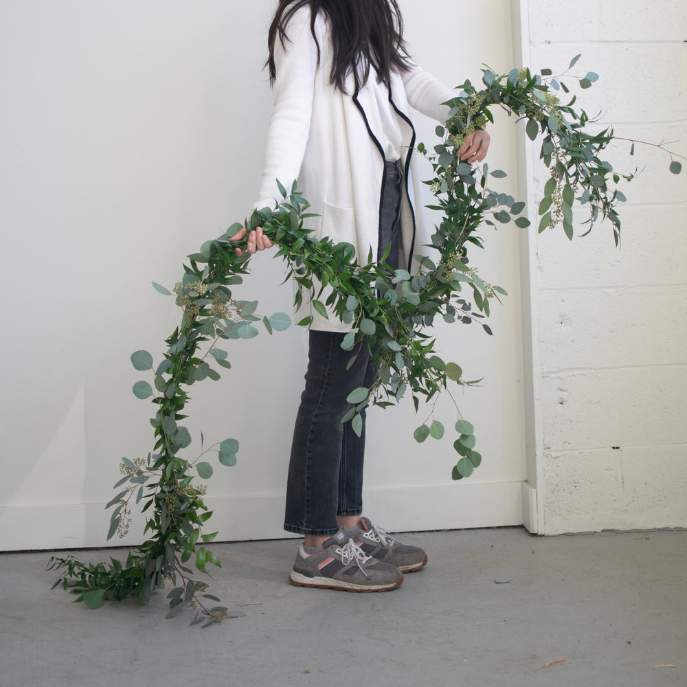 A woman in profile wearing a fancy white coat holds out a length of woven fresh greenery.