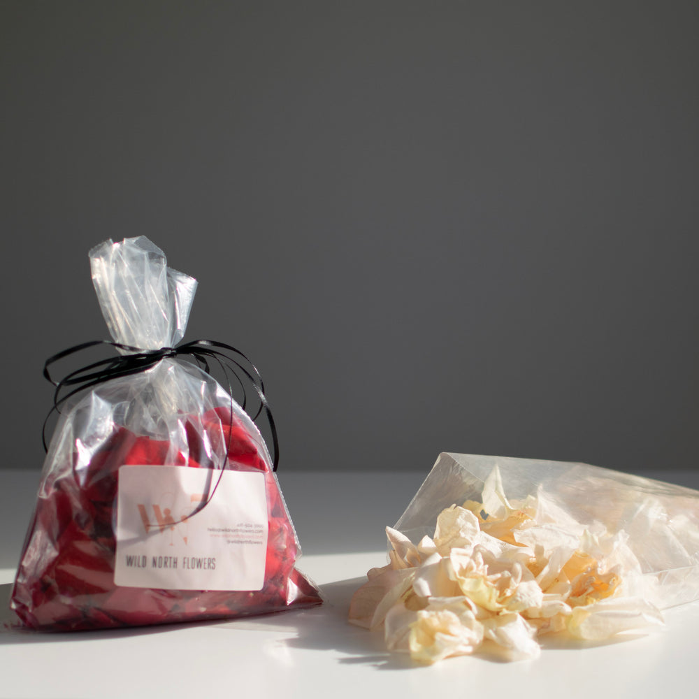 A bag of red rose petals is tied up with black ribbon. To the right, white petals spill out of a bag.