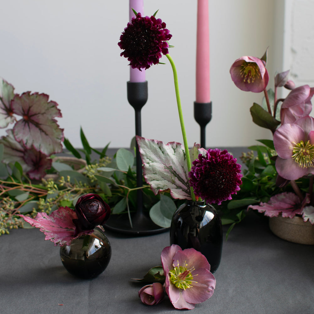 Deep purple carnations arranged in glossy black bud vases. Tapered candles and greenery can be seen in the background.