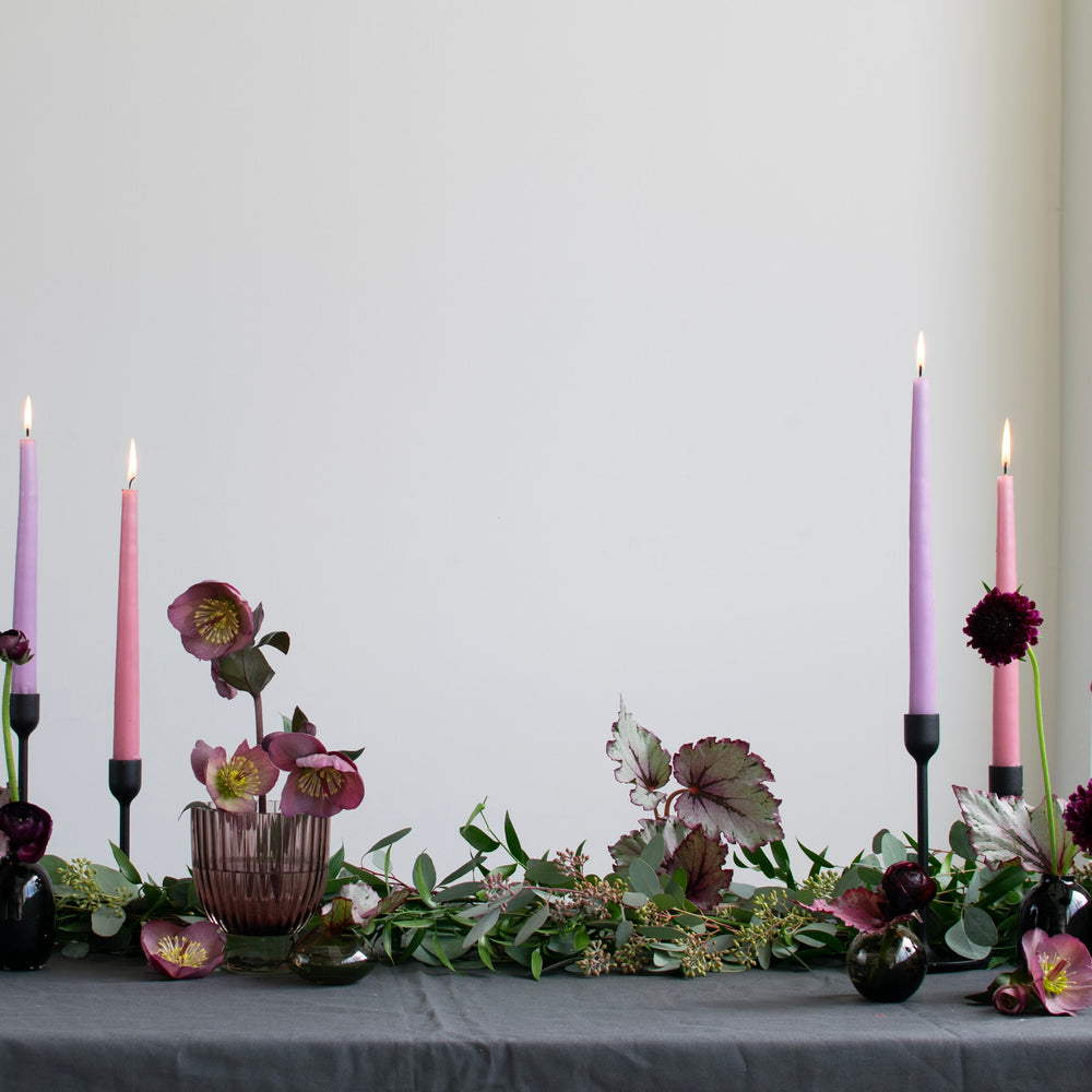 A greenery garland with pops of purple sits atop a tablecloth with simple flower blooms and candles.
