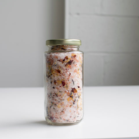 A glass jar with a metal cap contains pink himalayan bath salts and dried floral elements.
