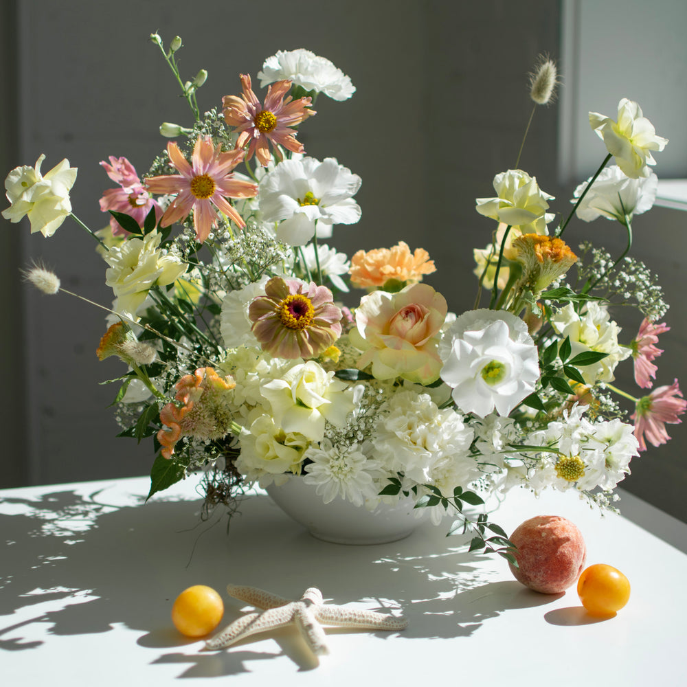 A white and cream floral arrangement with  muted pink accents sits on a table next to fresh fruits.