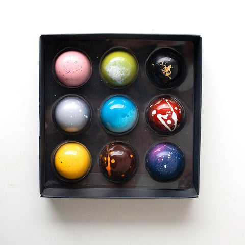 Nine chocolate truffles in a black box, made to look like a galaxy of planets in space.