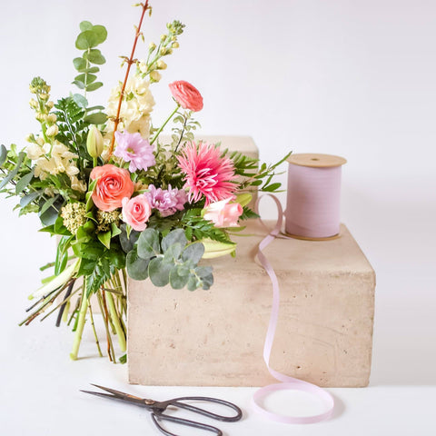 A hand tied bouquet sits on a concrete block with a spool of pink ribbon.