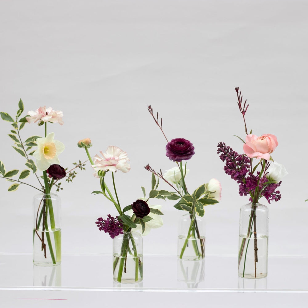 Flowers in deep purple and whites, including ranunculus and lilies, are displayed in glass bud vases.