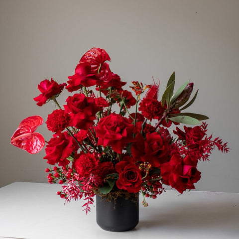 A large, all red floral arrangement in a moody black vase.