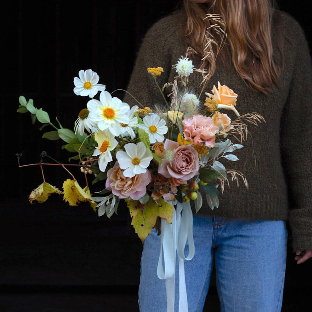 A woman wearing a sweater and blue jeans holds a fresh floral bouquet containing daisies and roses.