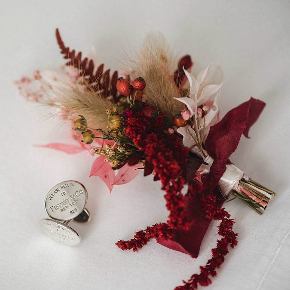 A moody burgundy boutonniere with red trailing flowers sits next to silver Tiffany & Co. cufflinks.