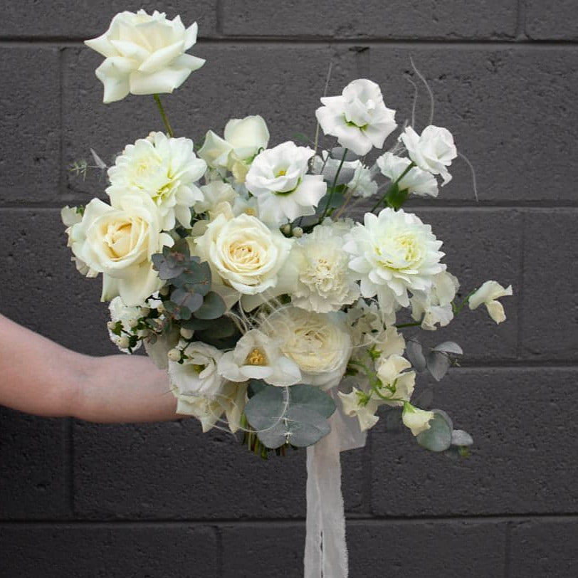 A classic, all white and cream bridal bouquet held against a grey brick wall.