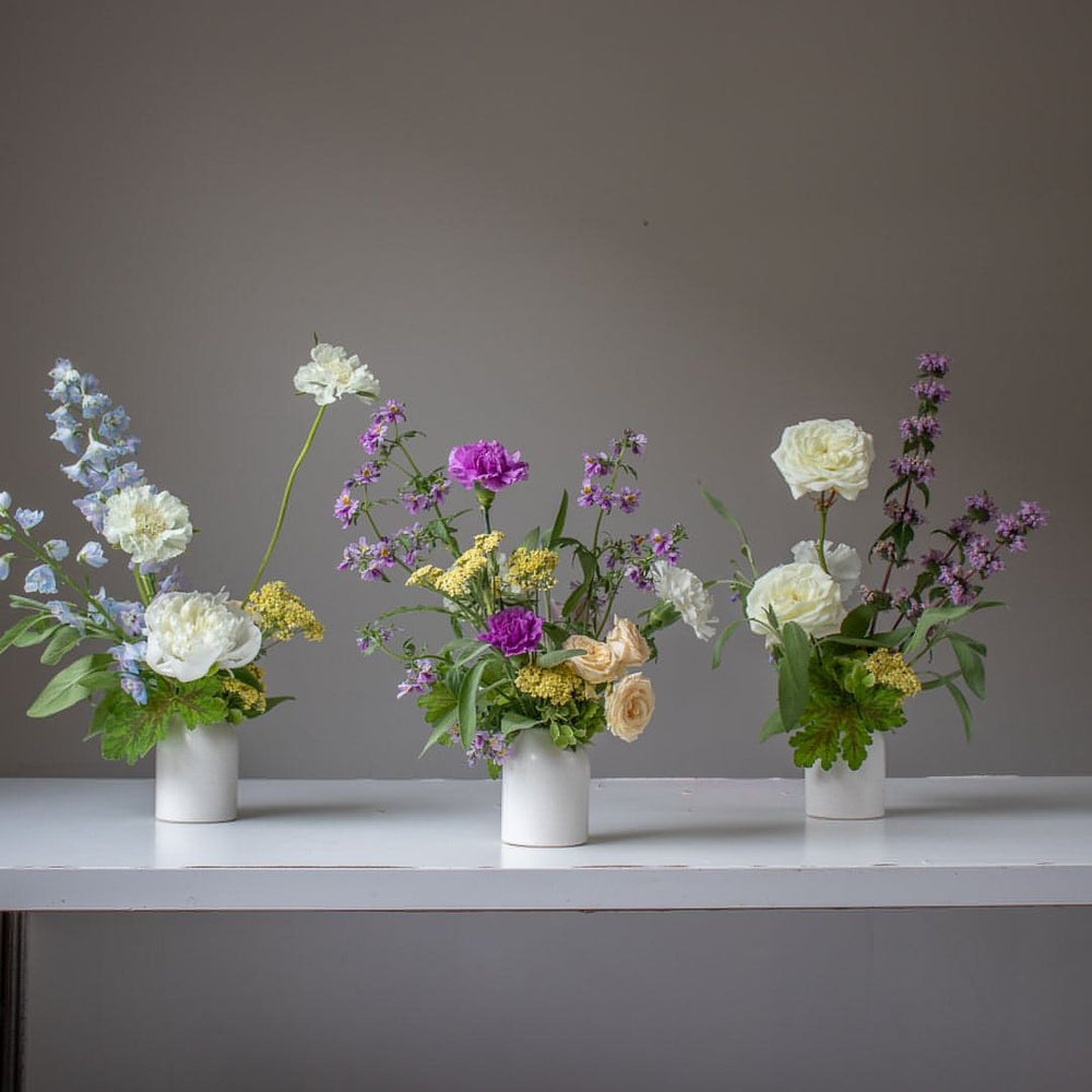 Three bud arrangements in mini cream vases. The palette includes deep purples, whites, soft blues, and yellows.