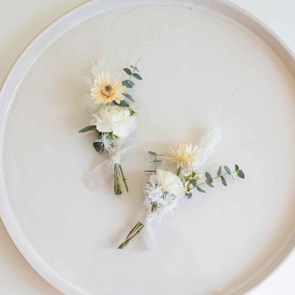 Two white and cream boutonnieres with eucalyptus stems are laid out on a round white plate.